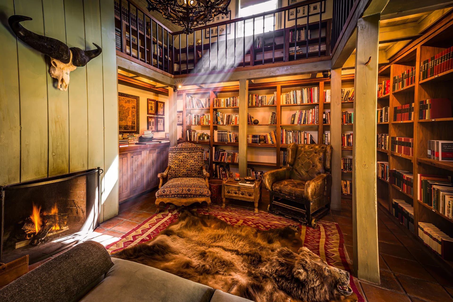 Cozy library with a fireplace on the left, a mounted bull skull, green paneled walls, two upholstered chairs, a fur rug, and tall bookshelves around a mezzanine loft.