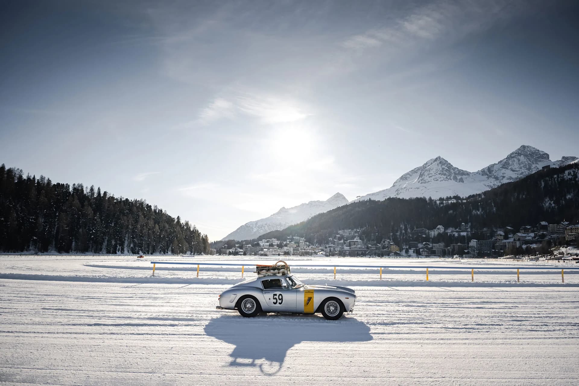 Snow-covered field with a silver vintage race car numbered 59; distant town, pine forest, and snow-capped mountains under a bright sun.