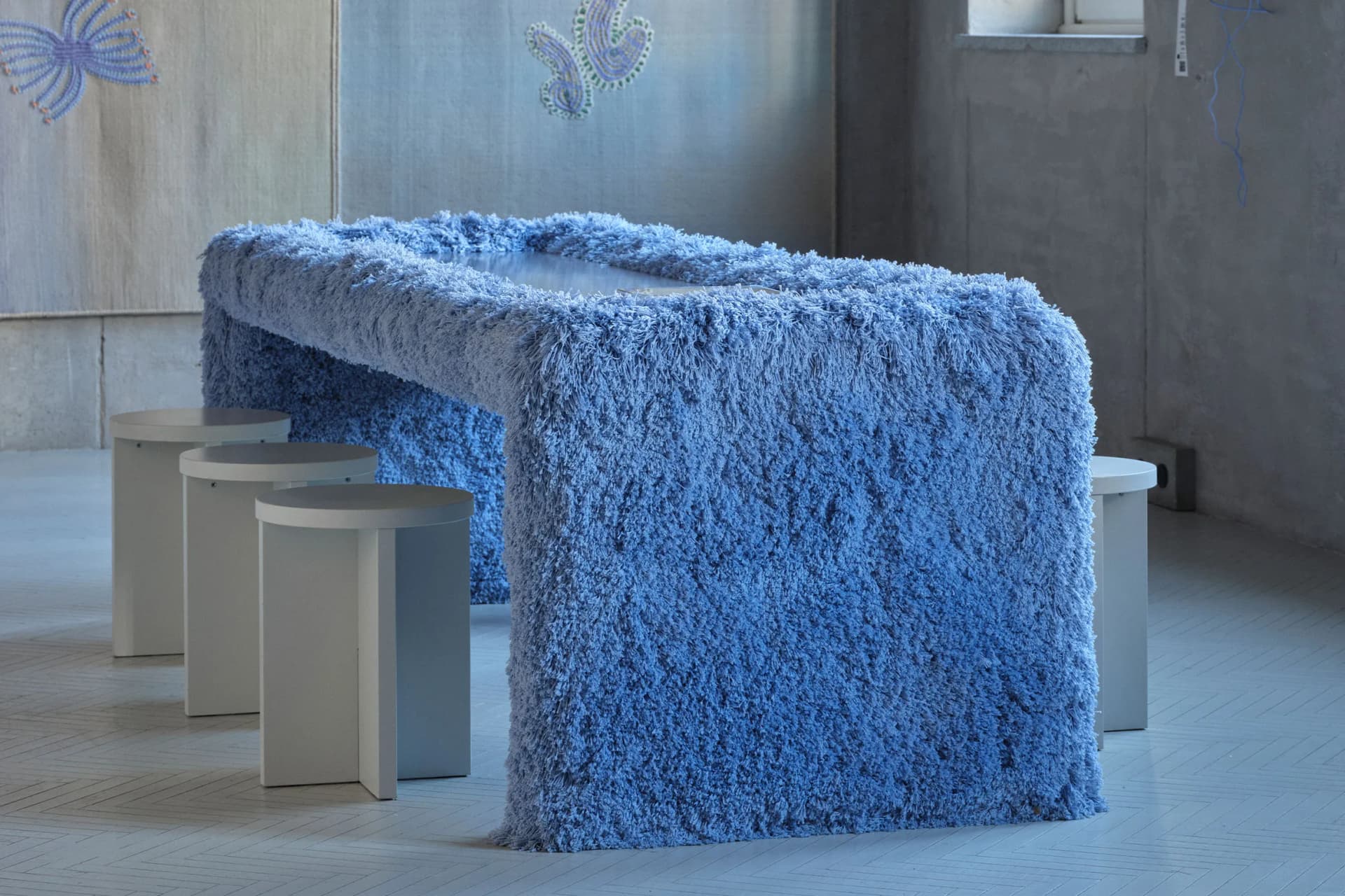 Large blue shag-covered table with round white stools in a minimalist room; gray walls and butterfly wall art.