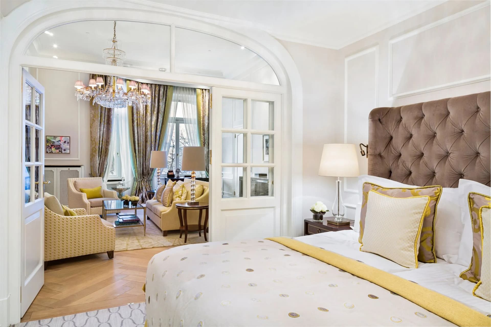 Una lussuosa camera da letto con una testiera imbottita color beige, biancheria da letto bianco-dorata e una zona soggiorno con un lampadario di cristallo.