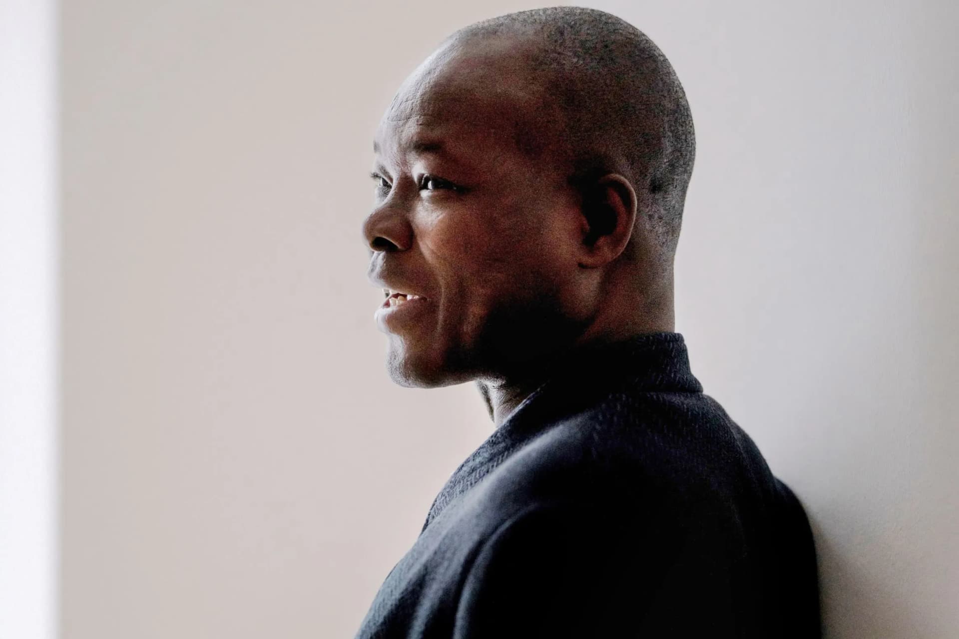 A profile shot of a smiling man with a shaved head, wearing a dark jumper, against a light, neutral background.