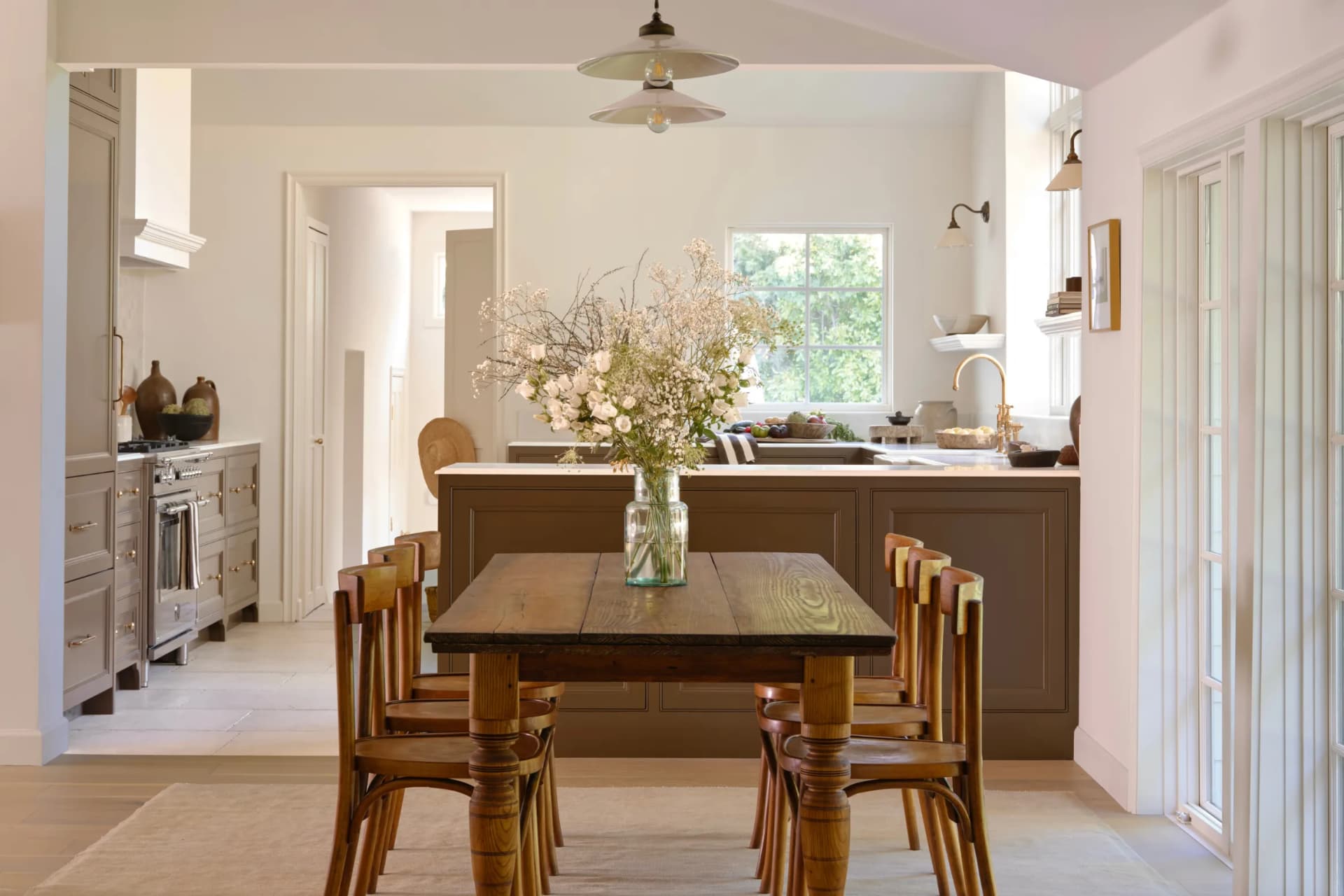 Bright, open kitchen and dining area with a wooden table and six chairs, a vase of white flowers on the table, and a dark island in the background.
