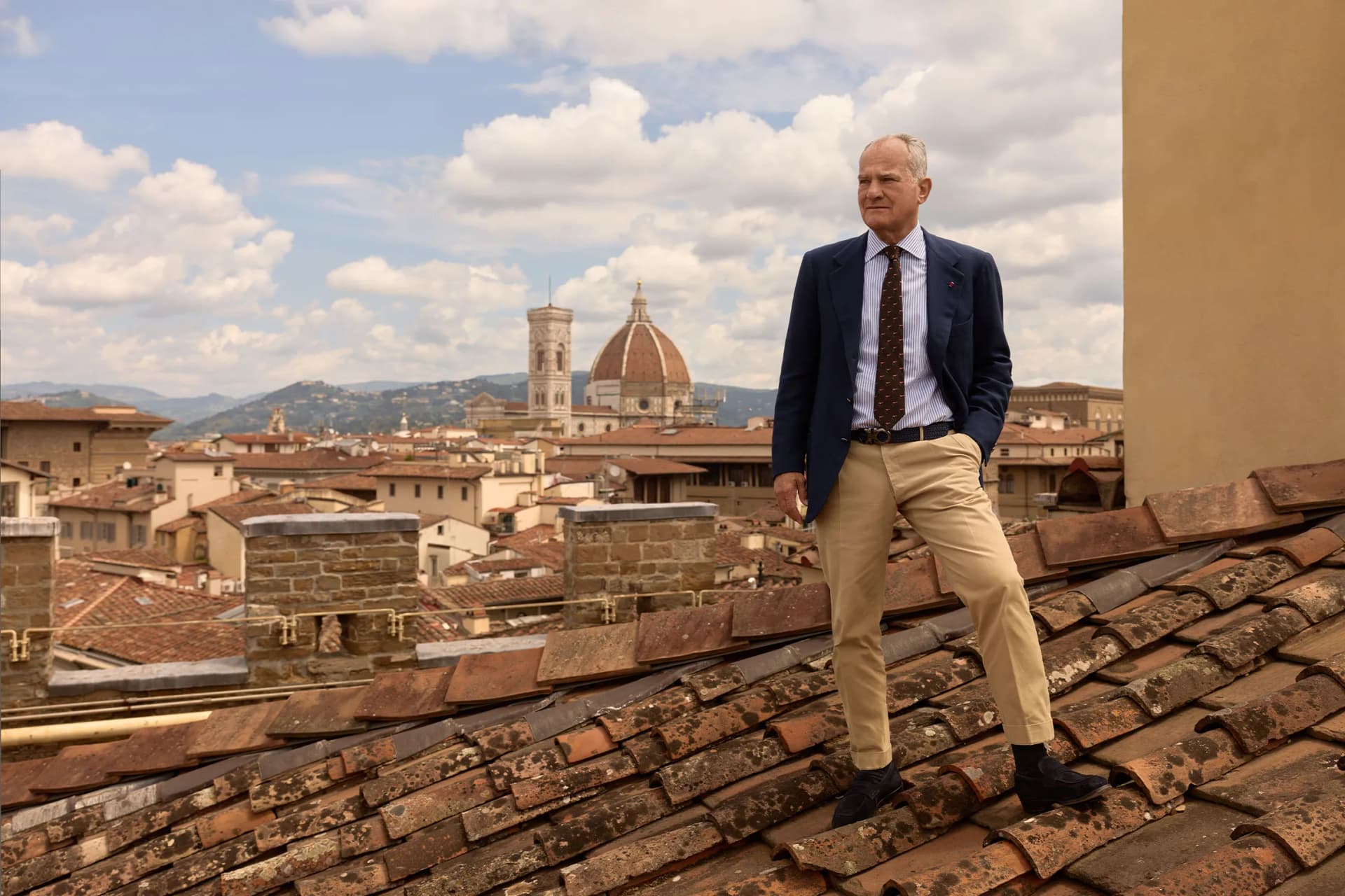 A man in a suit stands on a terracotta rooftop, overlooking a historic city with a domed cathedral and distant mountains.