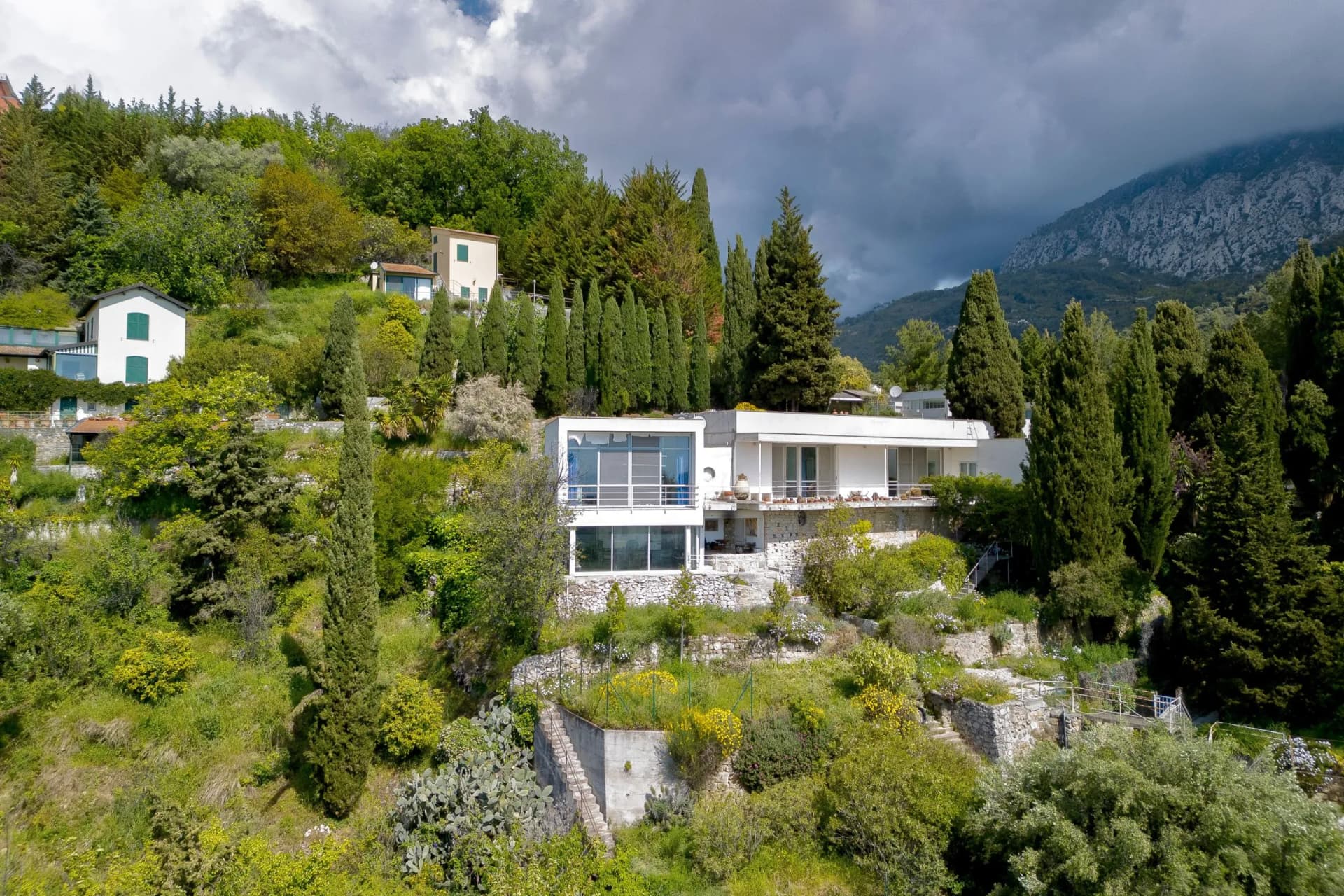 Hilly hillside lined with white modern homes among tall cypress trees, terraced gardens, and stormy clouds over distant mountains.