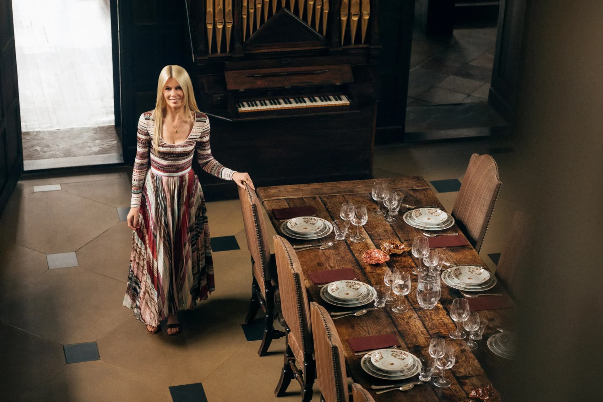 Blonde woman in a long striped dress stands by a rustic wooden dining table set for a meal, with glassware and bread, organ in background.