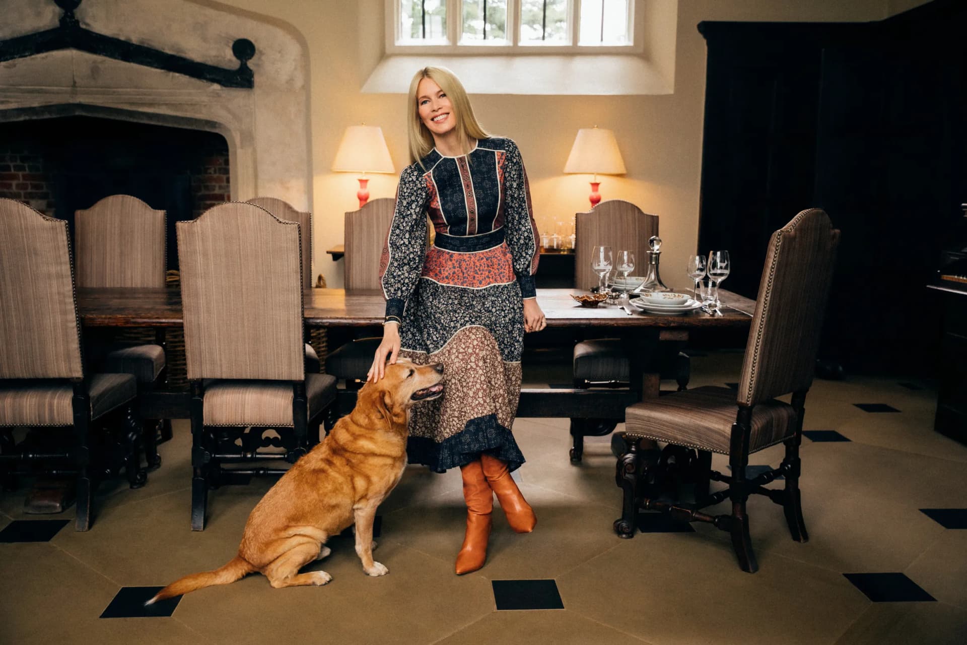 Smiling woman in a vintage patterned dress and orange boots stands with a golden retriever in a formal dining room with a table and stone fireplace.