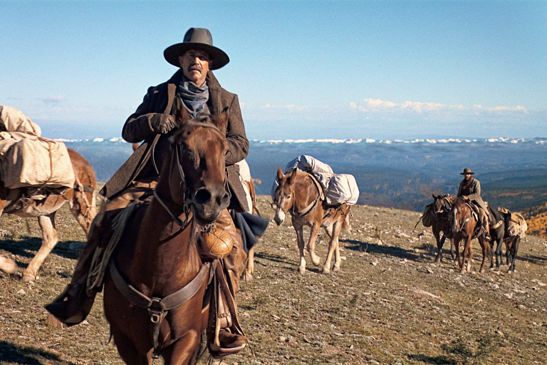 Cowboy with wide-brim hat rides a horse at the front as a line of riders and pack animals trek across a rocky hillside under a blue sky.