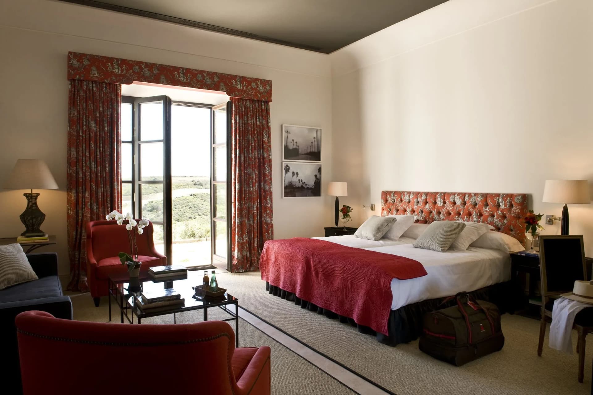 Hotel room with a single red tufted headboard bed, white linens, and red patterned curtains by tall windows; black-and-white wall art nearby.