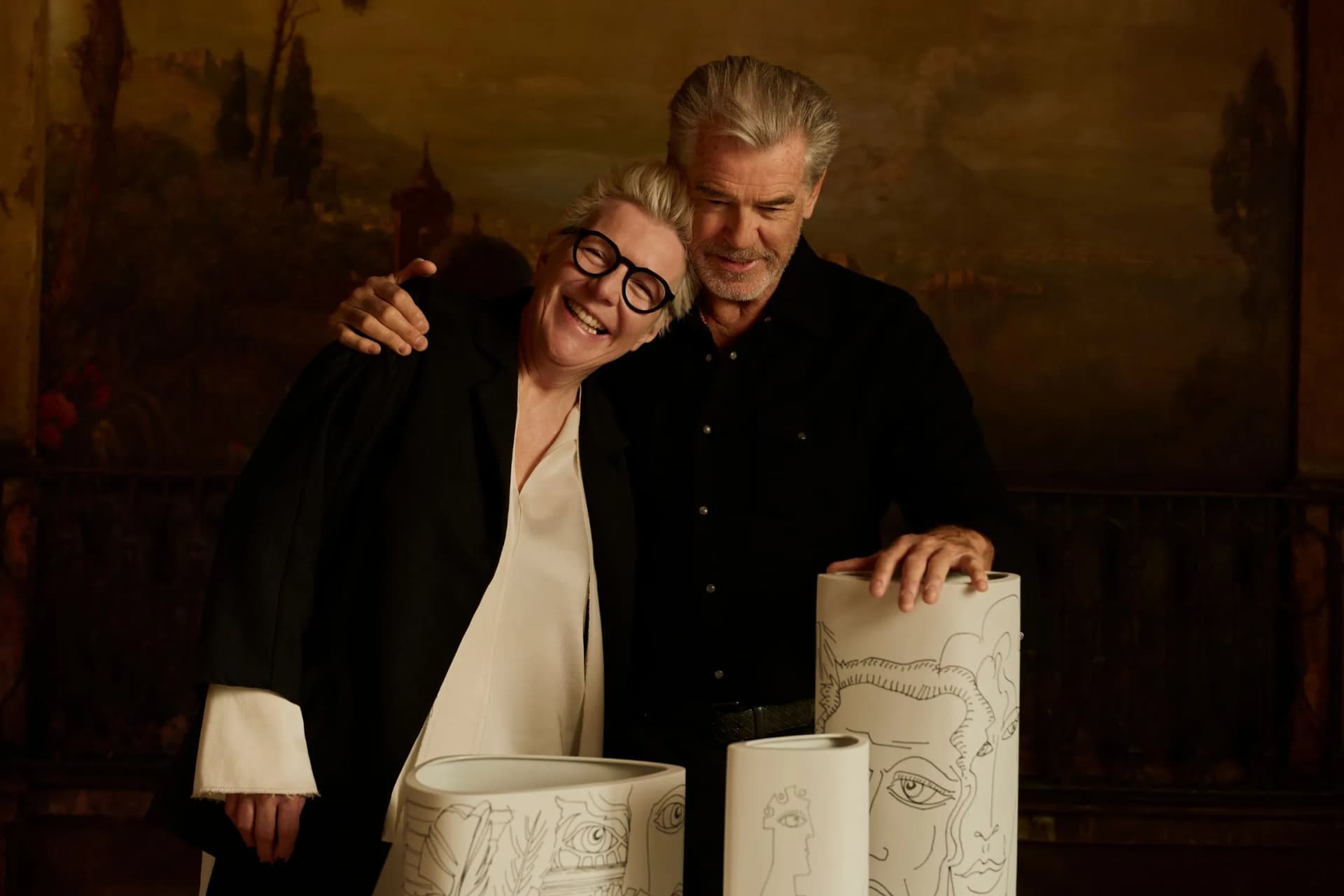 Two smiling adults stand close, arms around each other, beside cylindrical sculptures with line drawings in a warmly lit gallery.