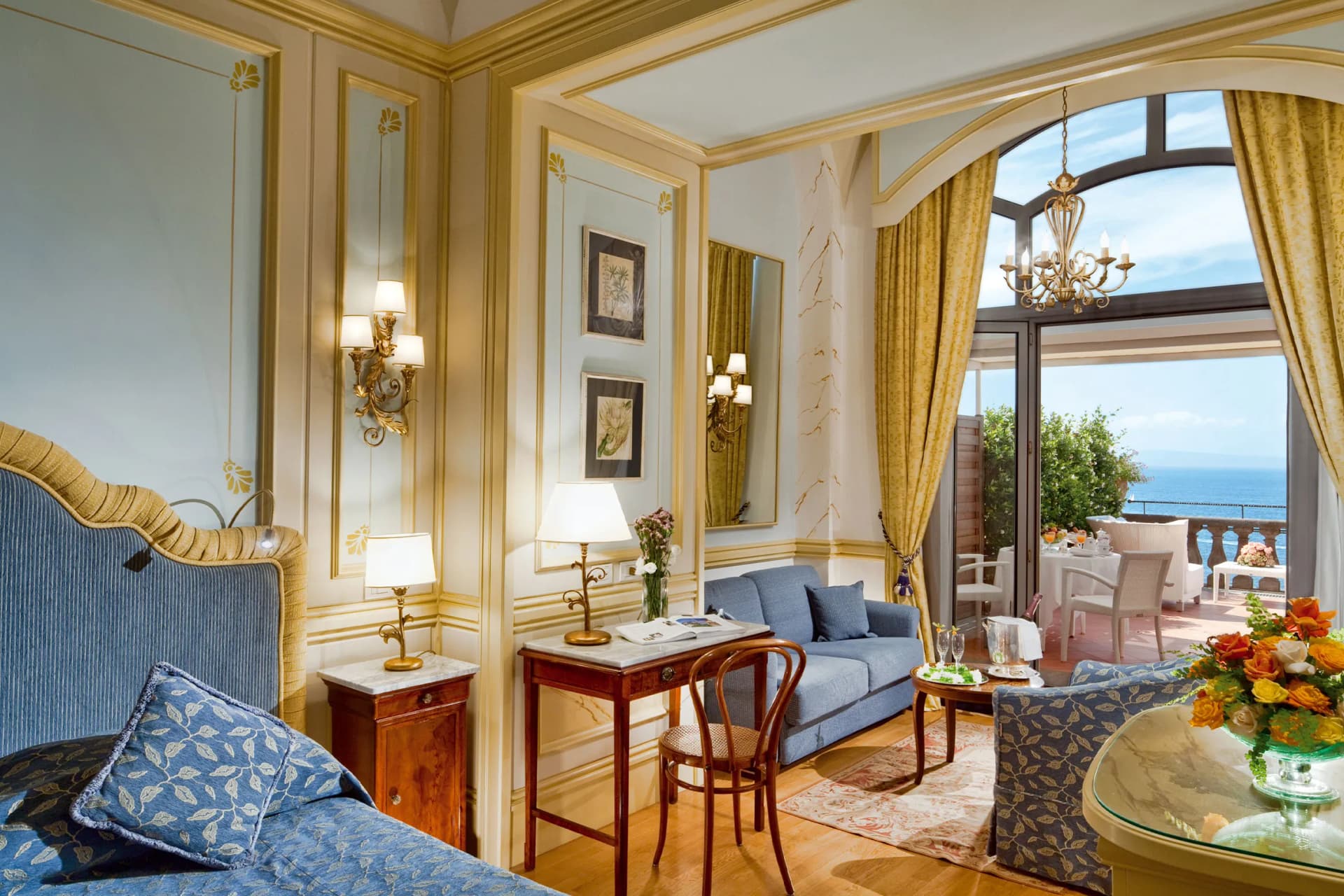 A luxurious living room with light blue walls, gold mouldings and a chandelier, opening onto a terrace with sea views.