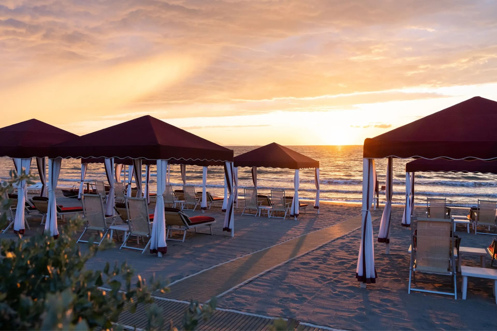 Sunset beach scene with burgundy cabanas and white curtains, rows of lounge chairs on the sandy shore facing the calm sea.