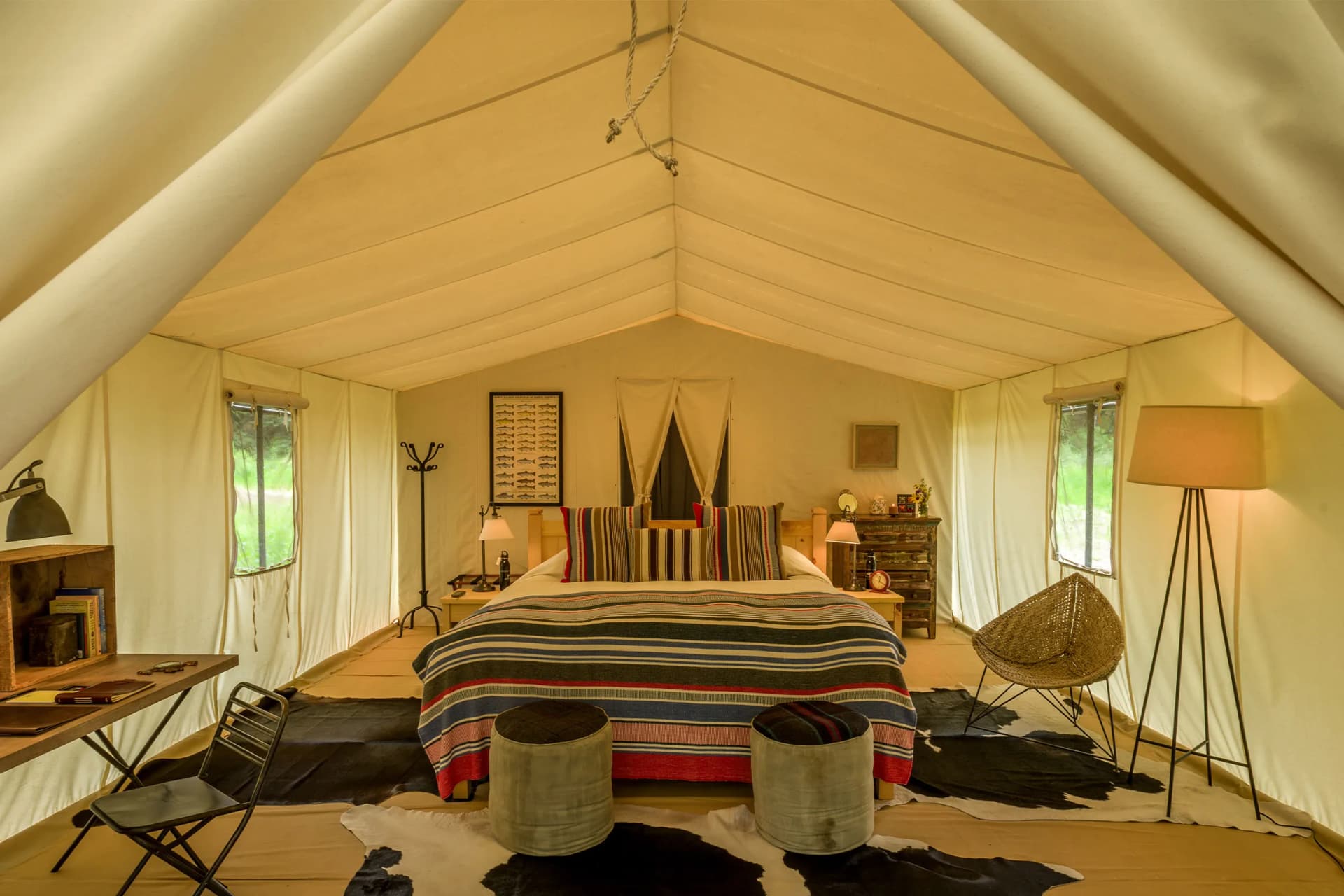 Inside a cream canvas tent bedroom with a striped bed, wooden furniture, two ottomans, a cowhide rug, and large windows.