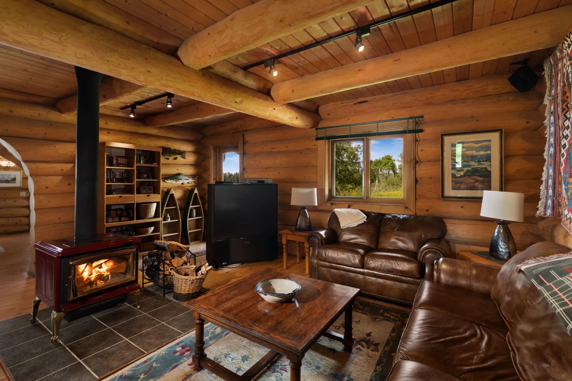 A cosy living room in a log cabin with exposed wooden beams, a wood-burning stove, a leather sofa, shelves and a window through which you can see the trees outside.