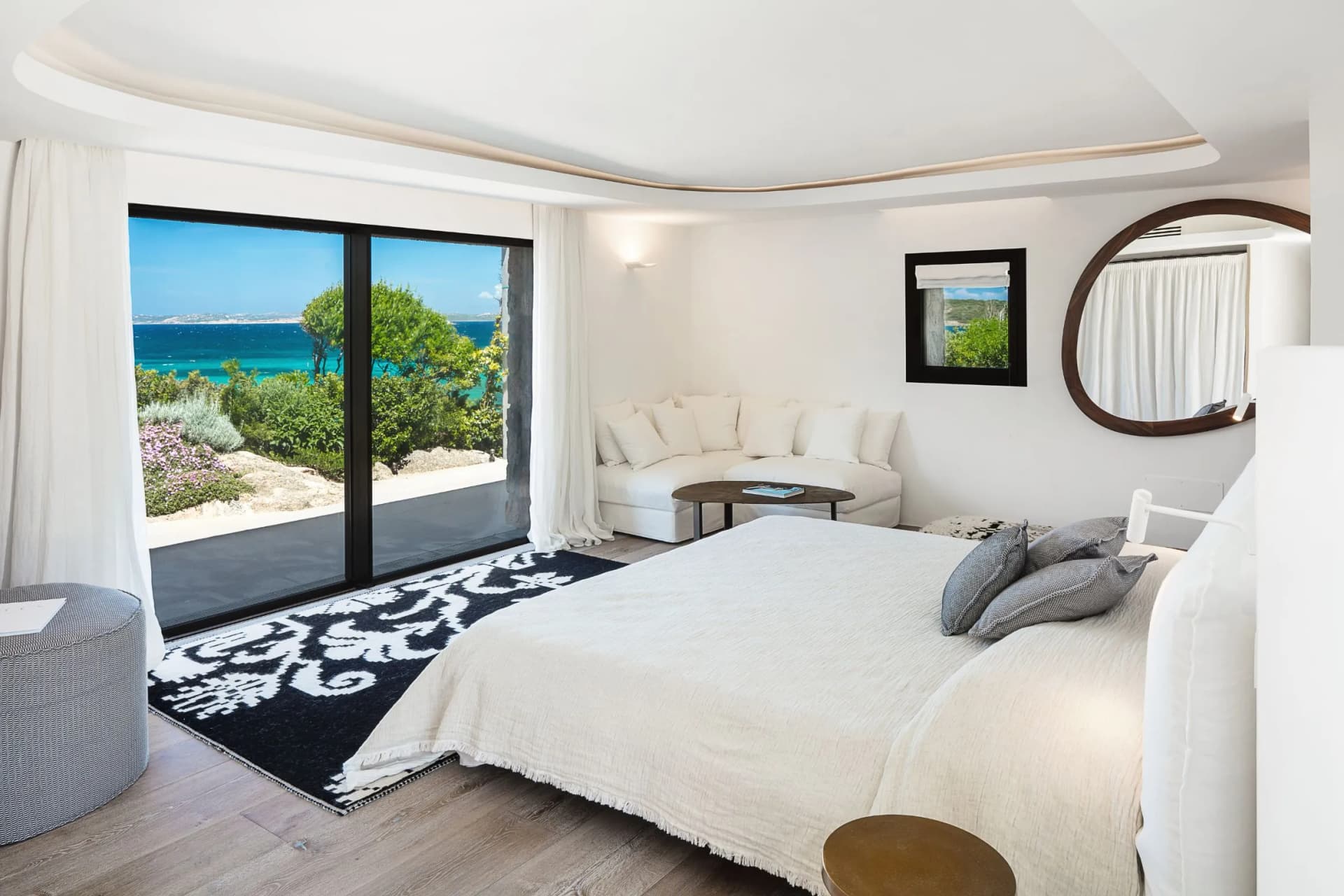 Bright coastal suite with all-white decor, large sliding glass door opening to the blue sea and greenery, a white sofa, and a round wooden-framed mirror.
