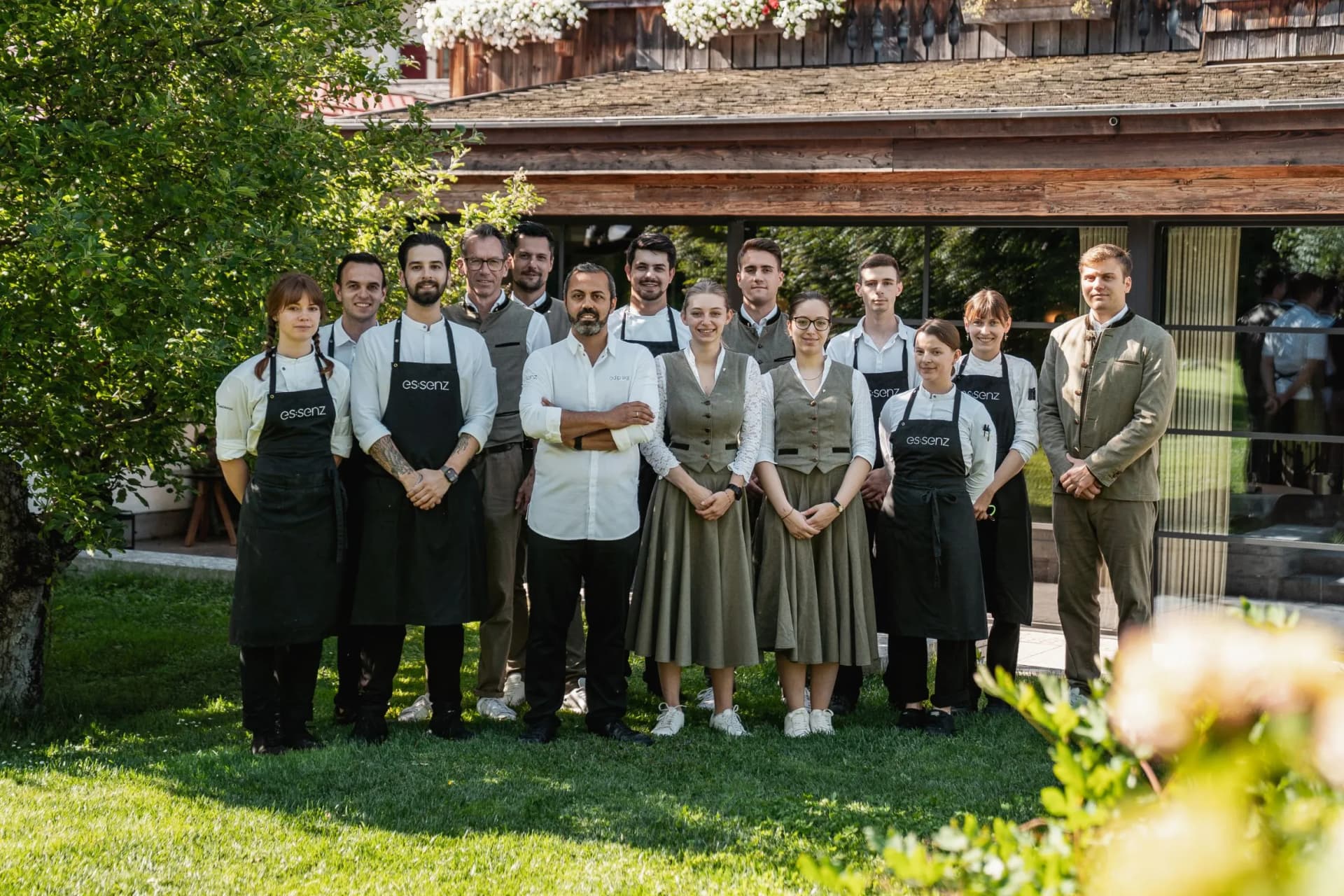 Group of restaurant staff in uniforms posing outdoors in front of a rustic wooden building, surrounded by greenery.