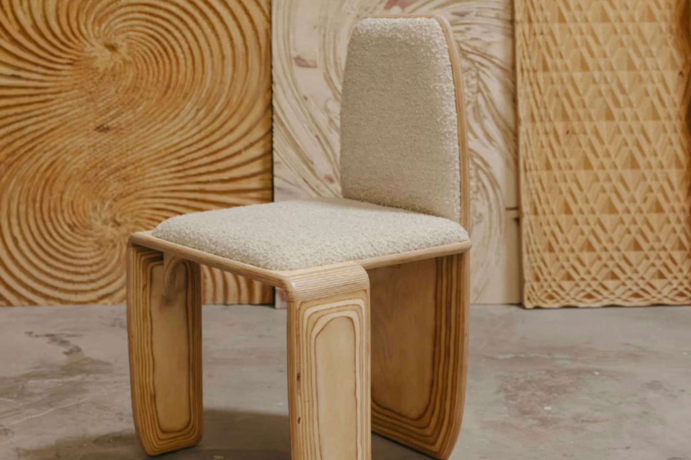 A pale wooden bent-plywood chair with a beige cushioned seat and backrest sits in a workshop with textured plywood panels behind.