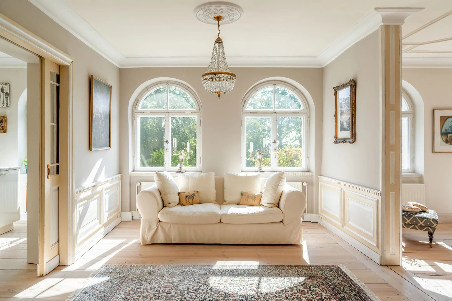 Elegant living room with cream walls, a grand crystal chandelier, arched windows, a beige sofa, framed art, and a patterned rug.
