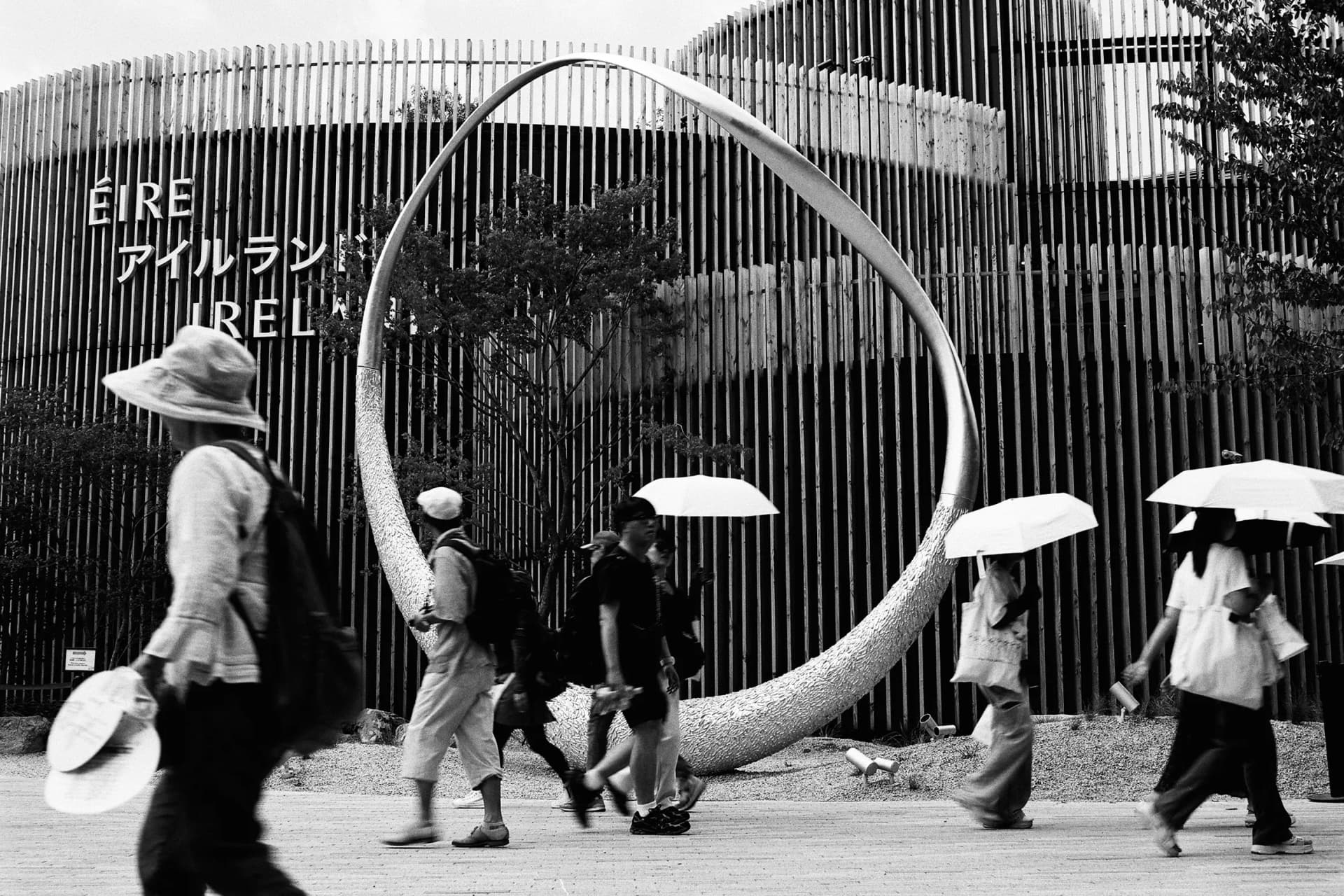 A black-and-white street scene: a huge, curved metal ring in front of a vertical wall of wooden slats bearing the inscription ‘EIRE’; pedestrians walk past carrying umbrellas.