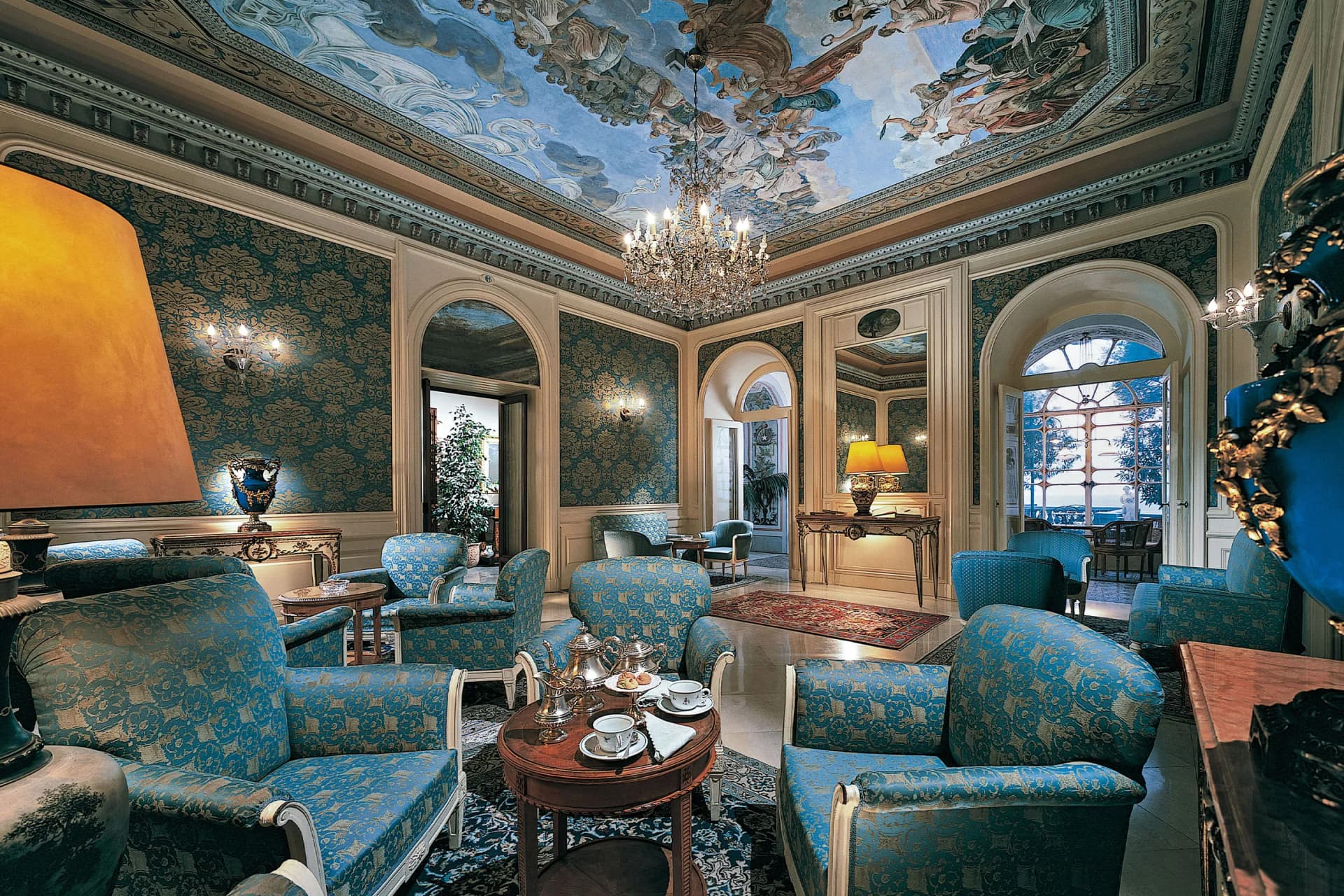 A sumptuous drawing room with turquoise damask-covered walls, an ornate ceiling fresco, a crystal chandelier and blue upholstered armchairs arranged around wooden tables.
