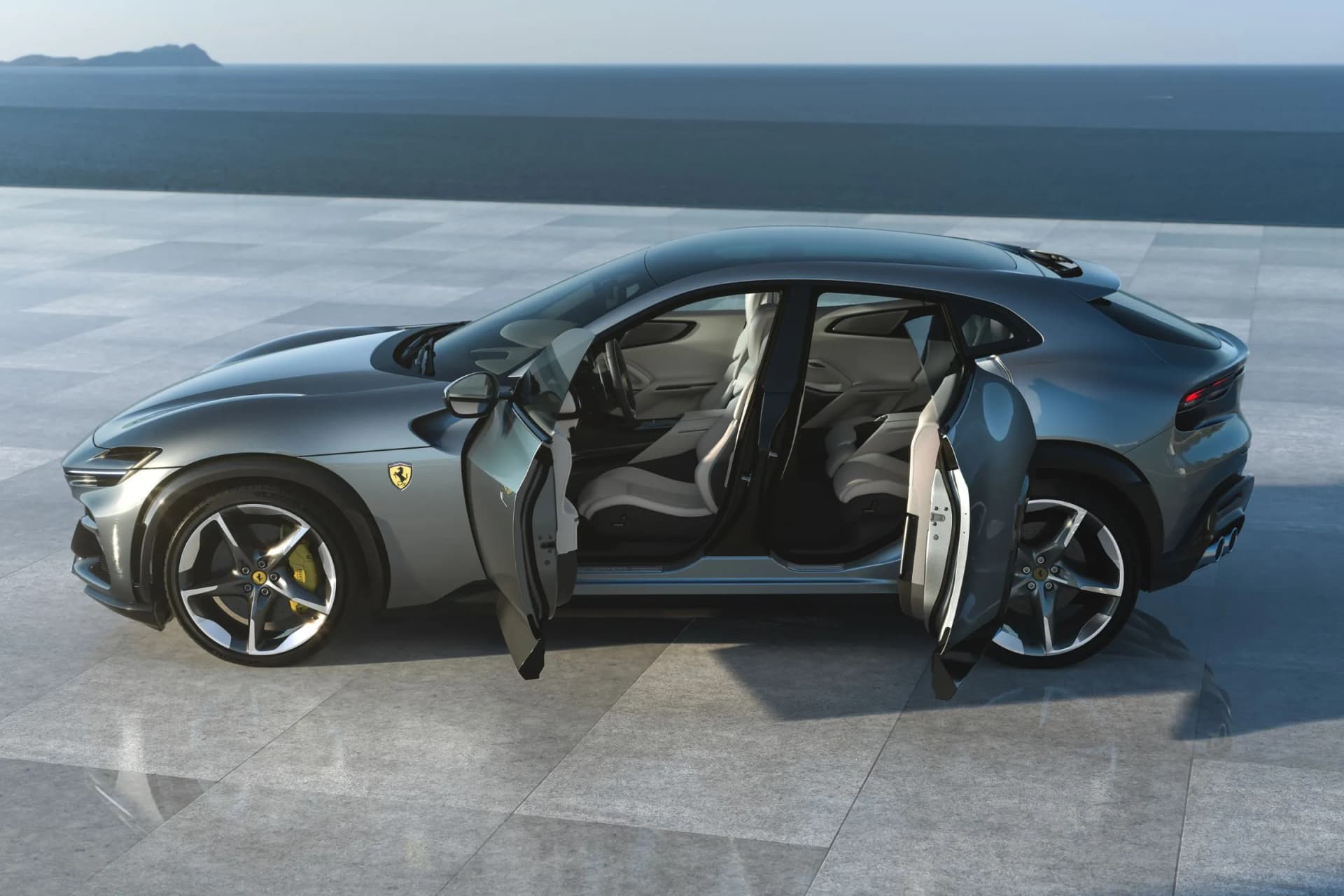 Two-door dark blue Ferrari with doors open, showing the leather interior on a tiled waterfront plaza with the sea in the background.