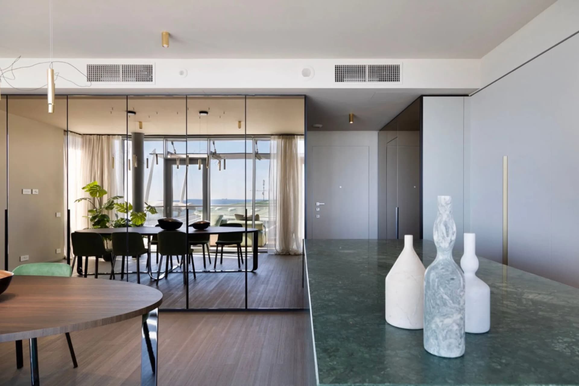 An open-plan living and dining area with glass partitions, a green marble kitchen island featuring ceramic vases, and large windows with sheer curtains