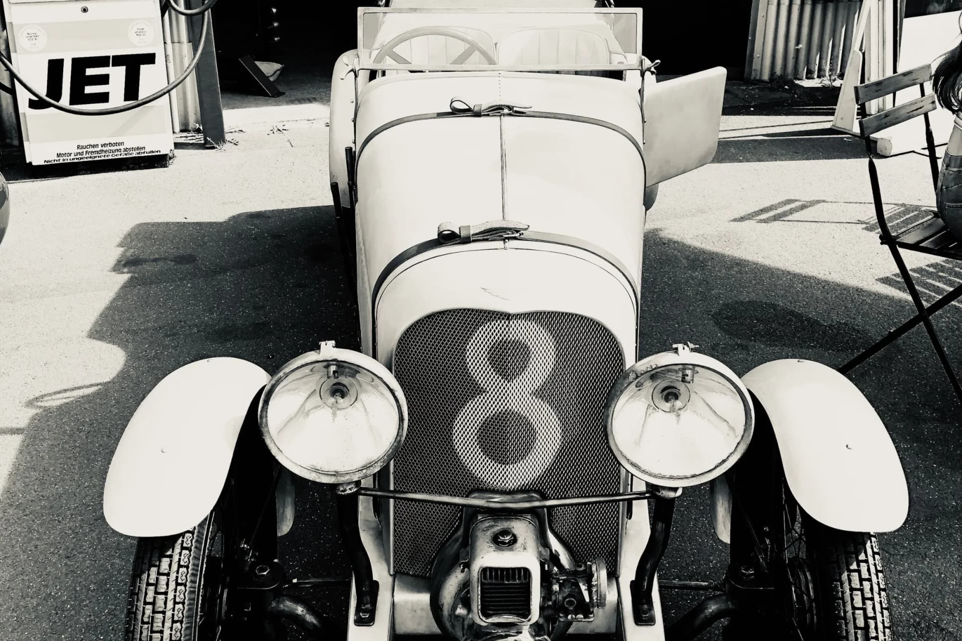Front view of a vintage race car with the number 8 on the grille, round headlights, and wide fenders in a pit area.