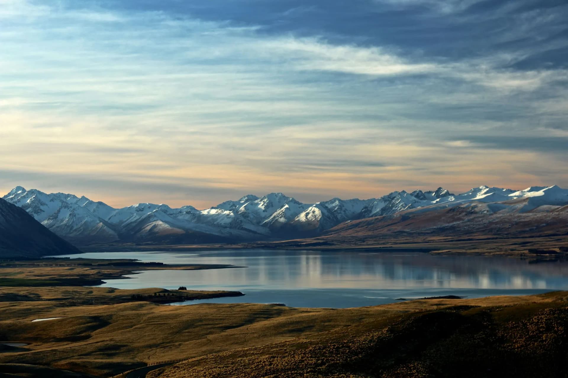 Snow-capped mountains reflect in a calm lake, with rolling brown plains in the foreground and a pastel sunset sky.