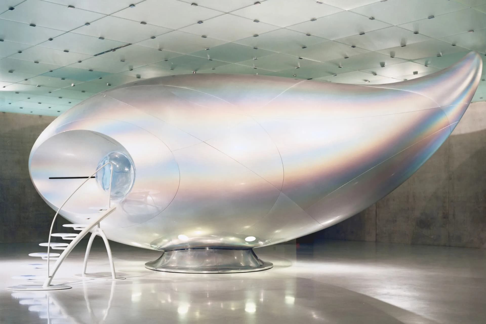 Large, polished silver teardrop capsule on a circular pedestal in a modern gallery, with a round viewing window at the front.