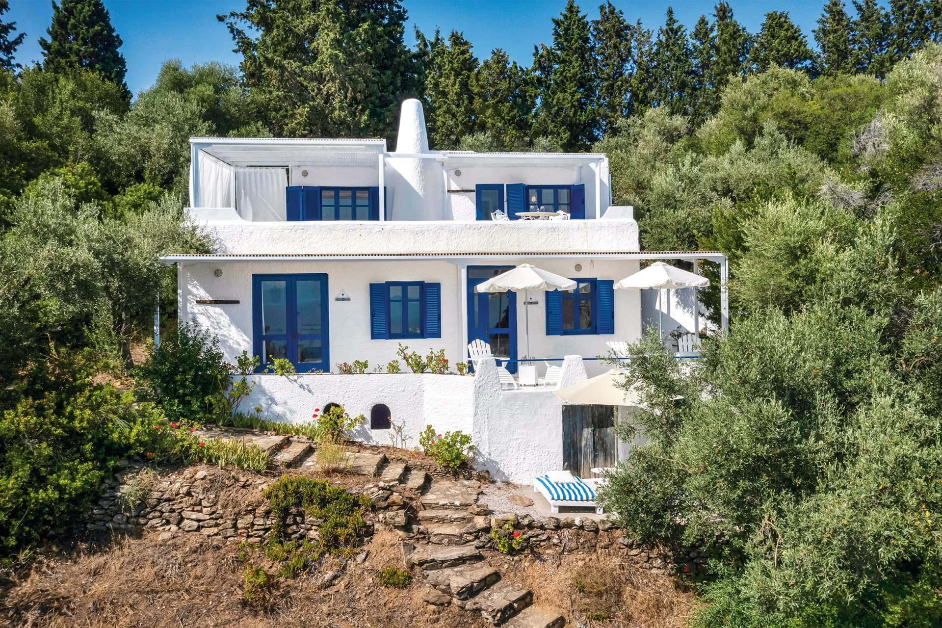 White-washed house with blue shutters on a rocky hillside, surrounded by dense greenery; outdoor patio with umbrellas.