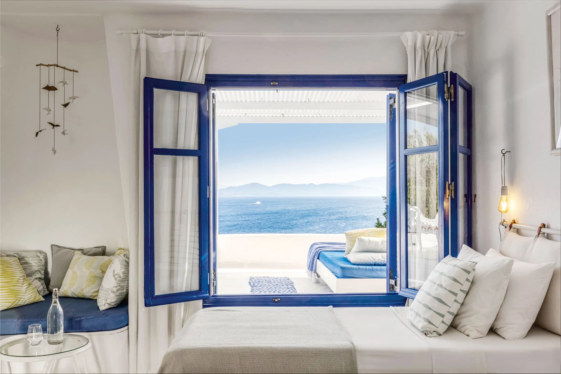 Bright seaside room with blue-framed doors opening to a balcony over the sea; white curtains, striped cushions, and a glass round table.