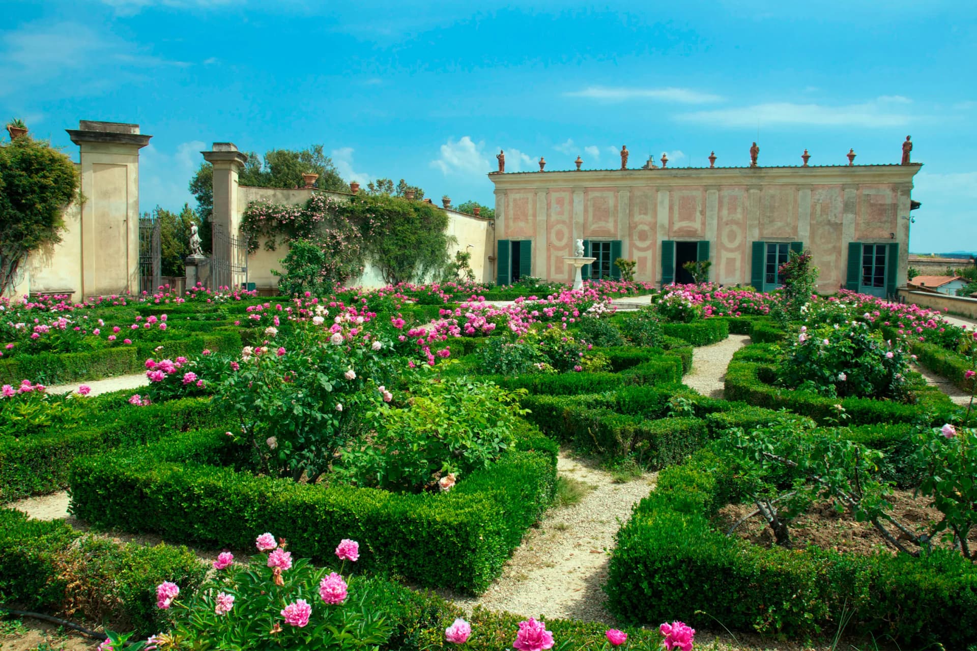 Sunlit formal rose garden with pink blooms and hedged paths, a pastel villa with green shutters, statues along the roof, and a central fountain.