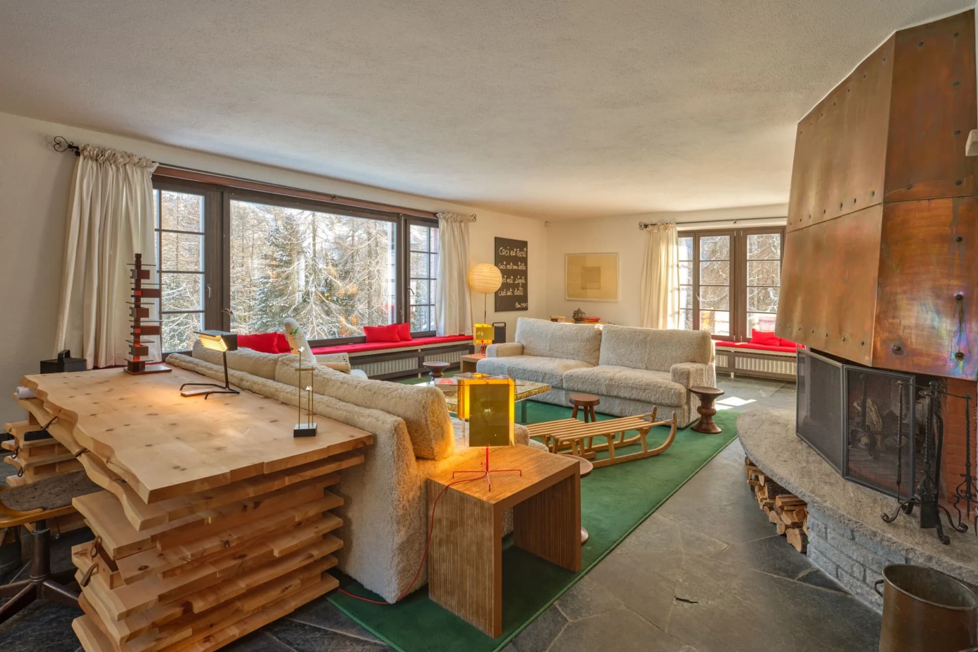 A cosy living room with large windows offering views of the snow-covered landscape, beige sofas, red cushions, wooden tables and a tall, rustic wooden fireplace