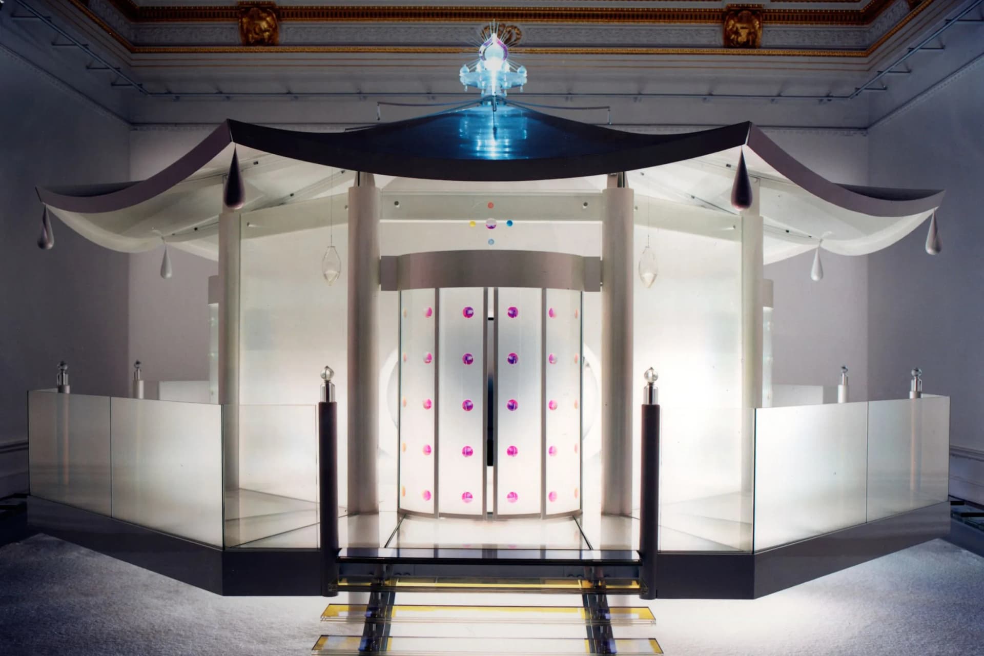 A glass pavilion with white panels and curved fabric drapes sits on a raised platform inside a grand room, topped by a blue glow and pink-dot wall.