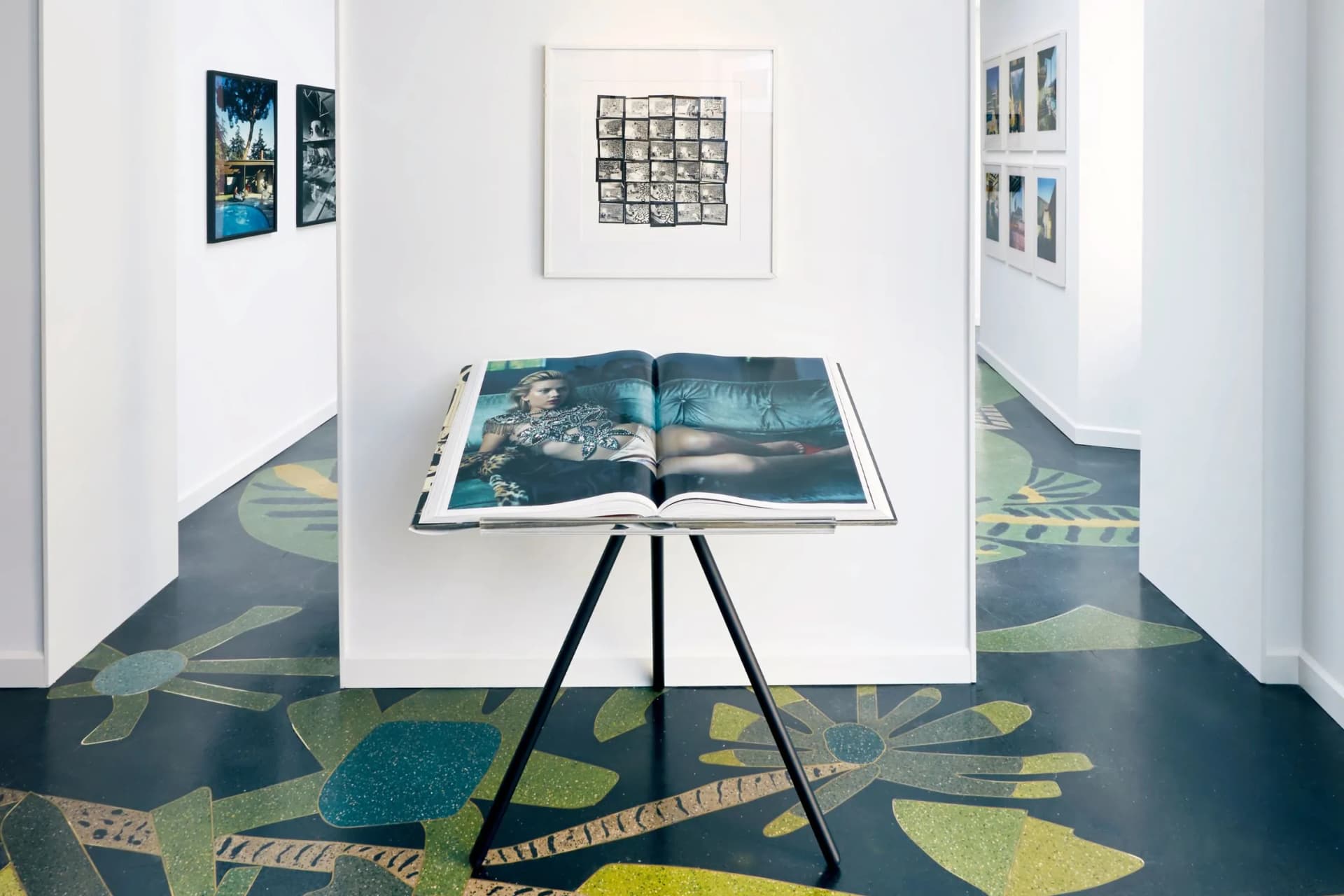Open art book on a tripod stand in a bright white gallery, with framed artworks on the walls and a colorful leaf-patterned floor.