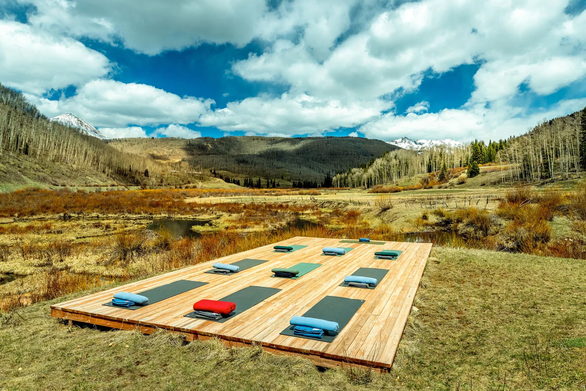 Outdoor wooden platform with yoga mats and cushions in a valley, trees lining the hills, snow-capped mountains under a bright blue sky with clouds.