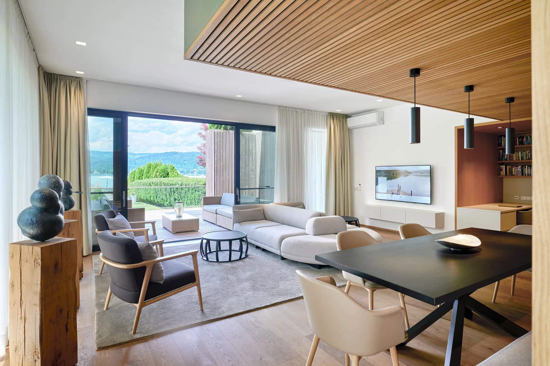 Bright living room with a wooden slat ceiling, large glass doors opening to a lake view, grey sofas, wooden chairs, and a wall-mounted TV.