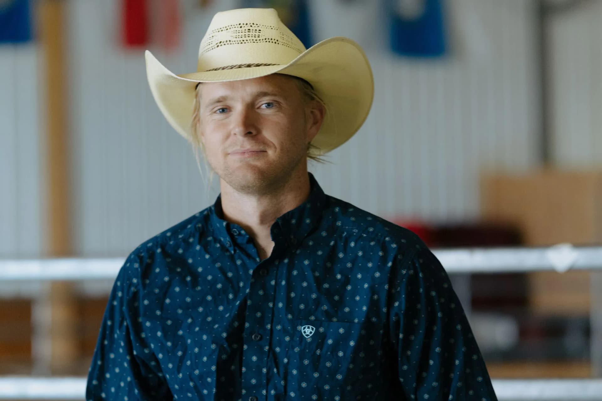 Blond man in a straw cowboy hat and blue patterned shirt, smiling softly in a blurred indoor setting.