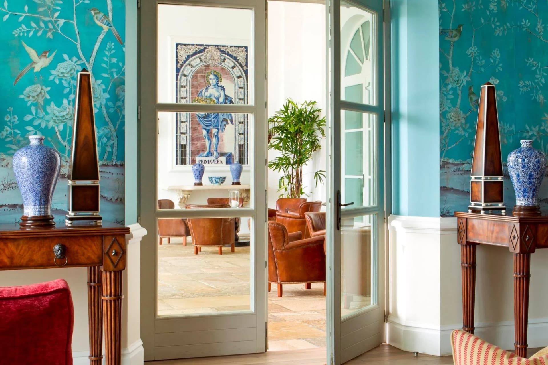 Teal wallpaper with birds and flowers, blue porcelain vases on wood consoles, a Primavera mosaic visible through glass doors, and a potted plant.