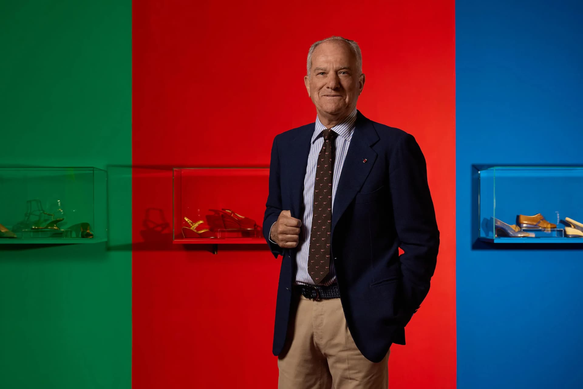 An older man in a navy blazer, white shirt, and khaki pants stands before a tri-color wall (green, red, blue) with glass display cases of shoes.