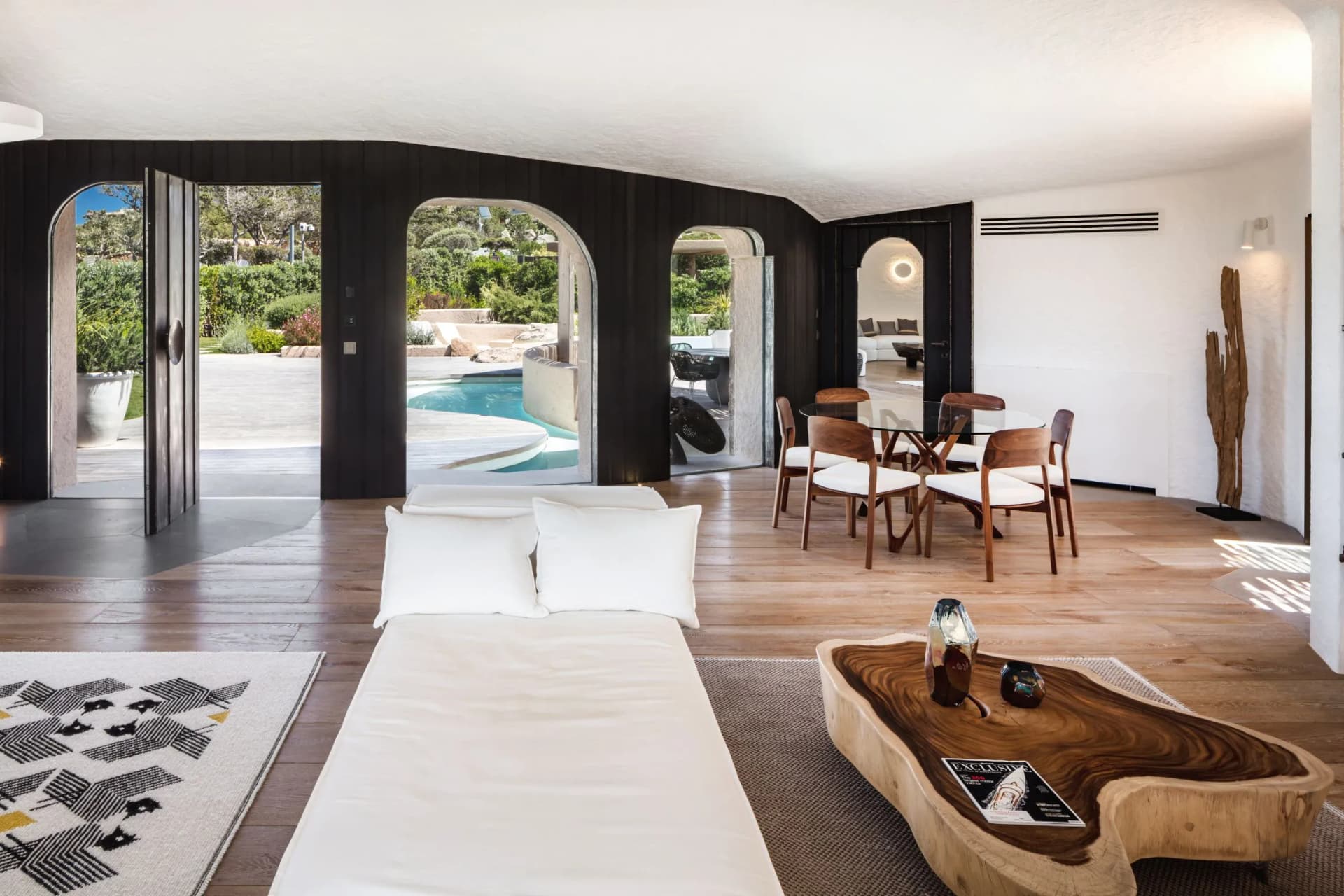 Open living space with dark curved walls and arched doorways opening to a pool and garden, white seating, and wooden furniture.