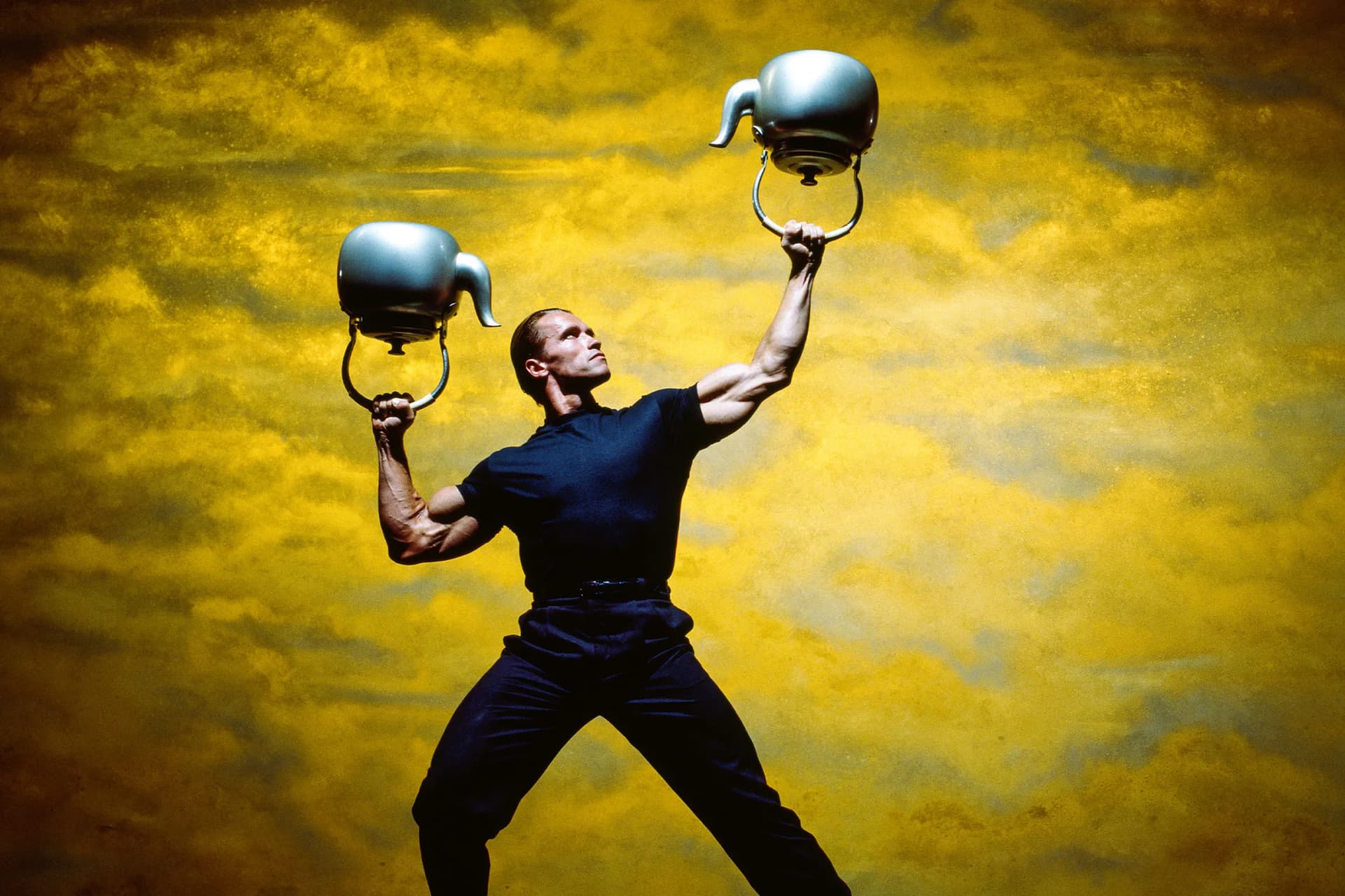 Muscular man in black athletic wear lifts two kettlebells overhead in a wide stance against a yellow cloudy sky.