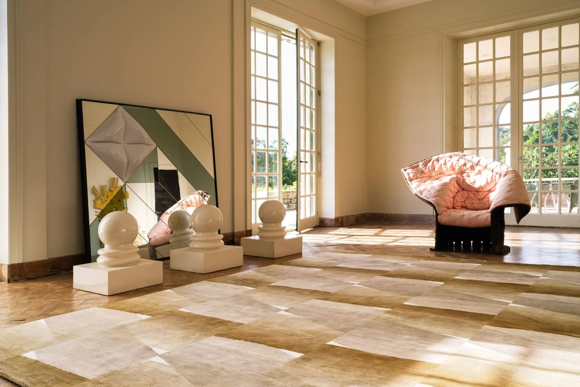 Bright room with large windows, a leaning geometric painting, white pedestal sculptures, and a pink-patterned armchair.