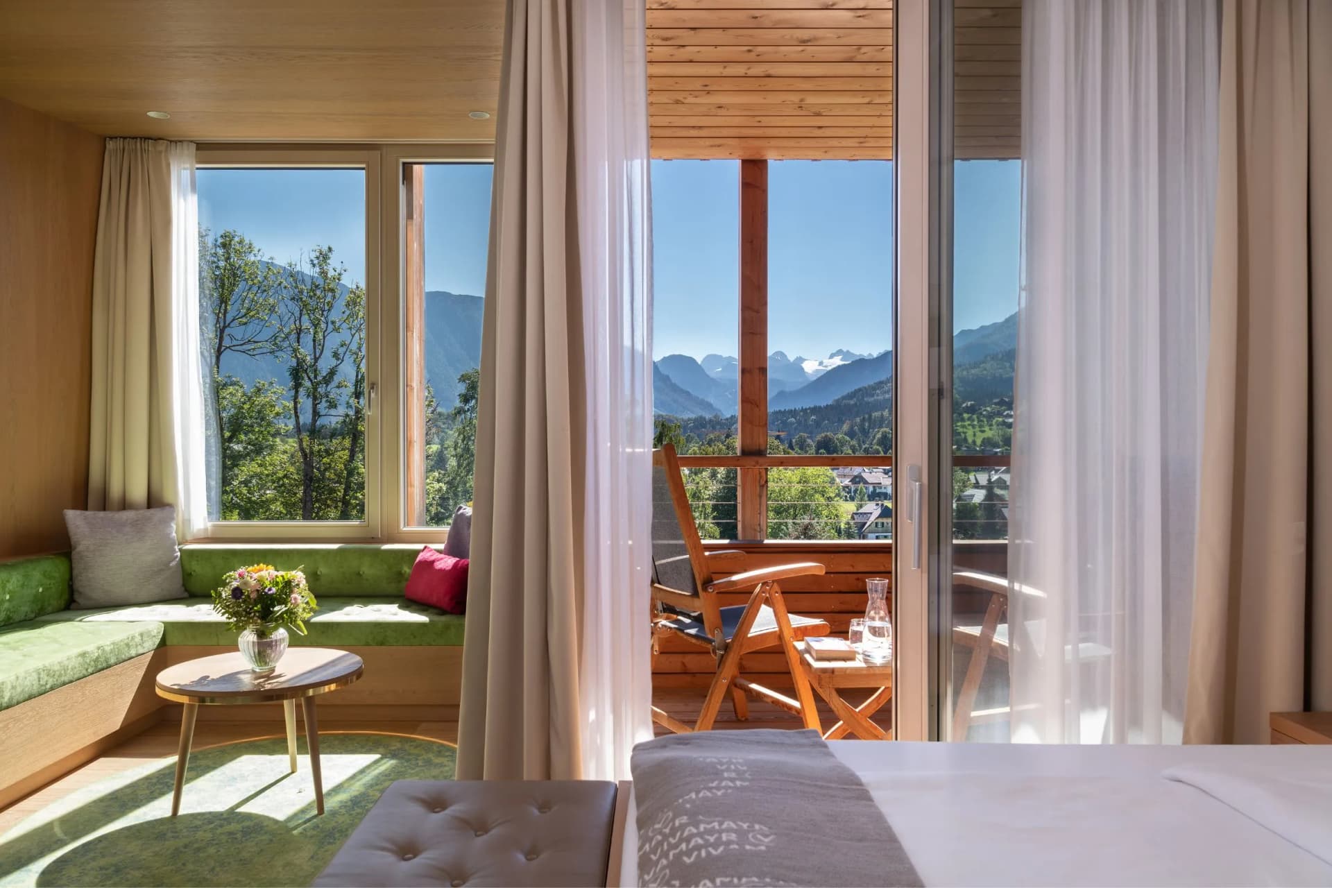 Bright hotel room with a green seating nook, a round coffee table with flowers, glass doors to a balcony with wooden chairs, mountain view.