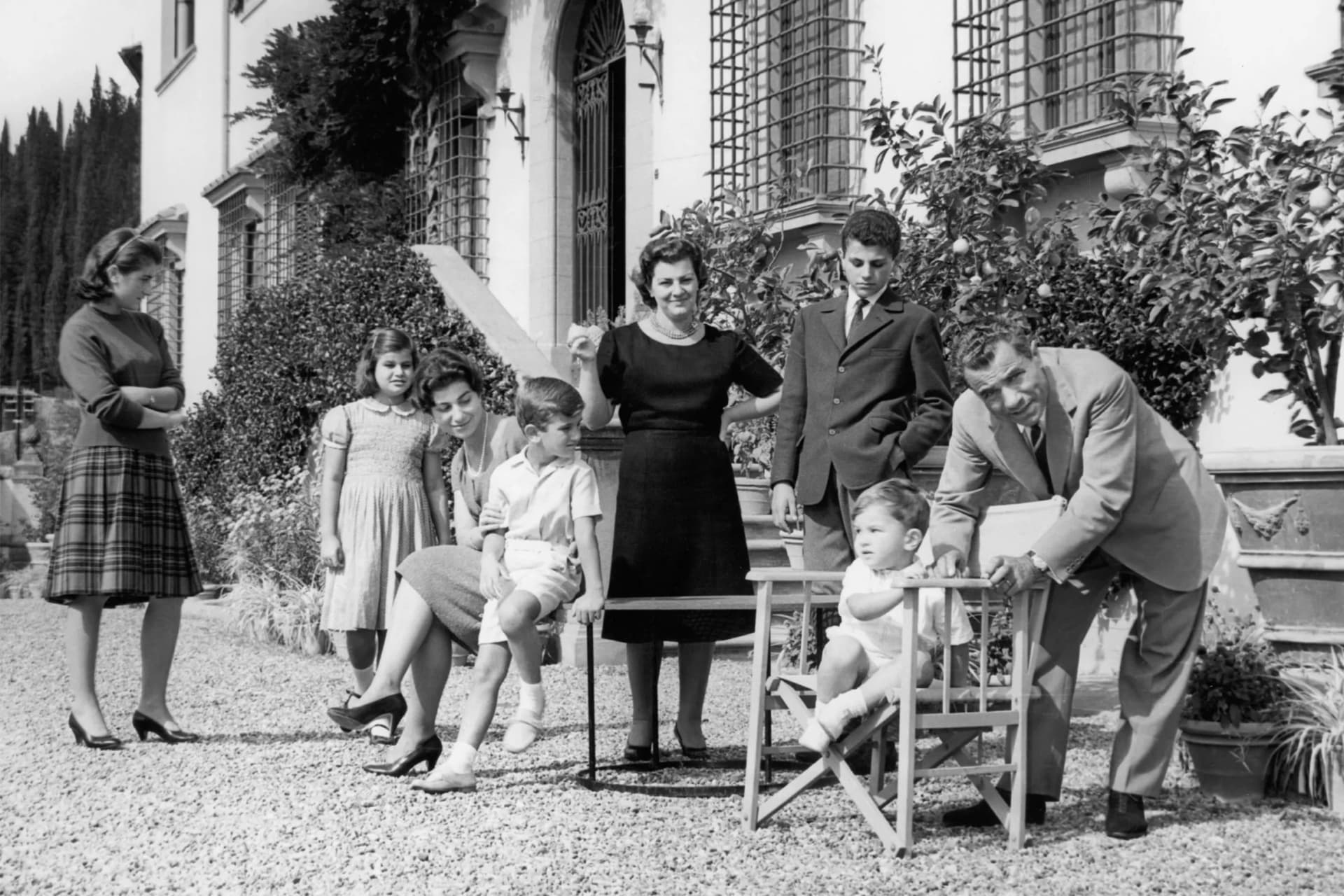 Black-and-white family photo outdoors in front of a villa with arched windows; adults and children pose on a gravel path as a man helps a toddler in a chair.