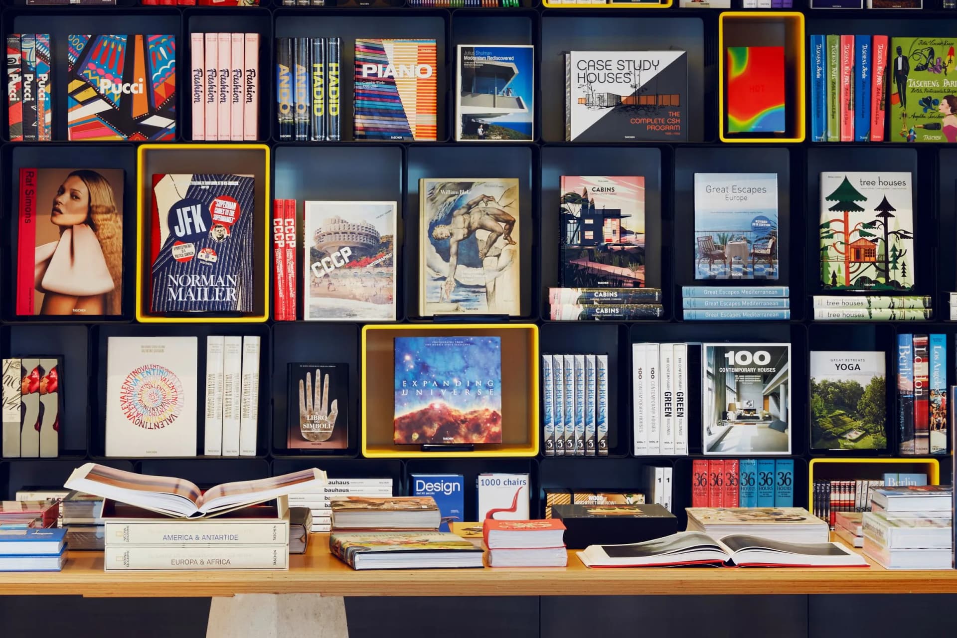 Colorful bookshelf wall filled with book covers in yellow-framed frames, and a wooden desk in front with open and stacked books.