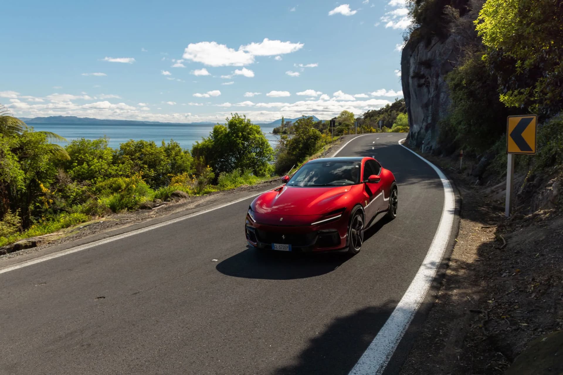 Red sports car on a winding coastal road with the sea on the left, a rocky cliff on the right, and a yellow curve sign ahead under a sunny blue sky.