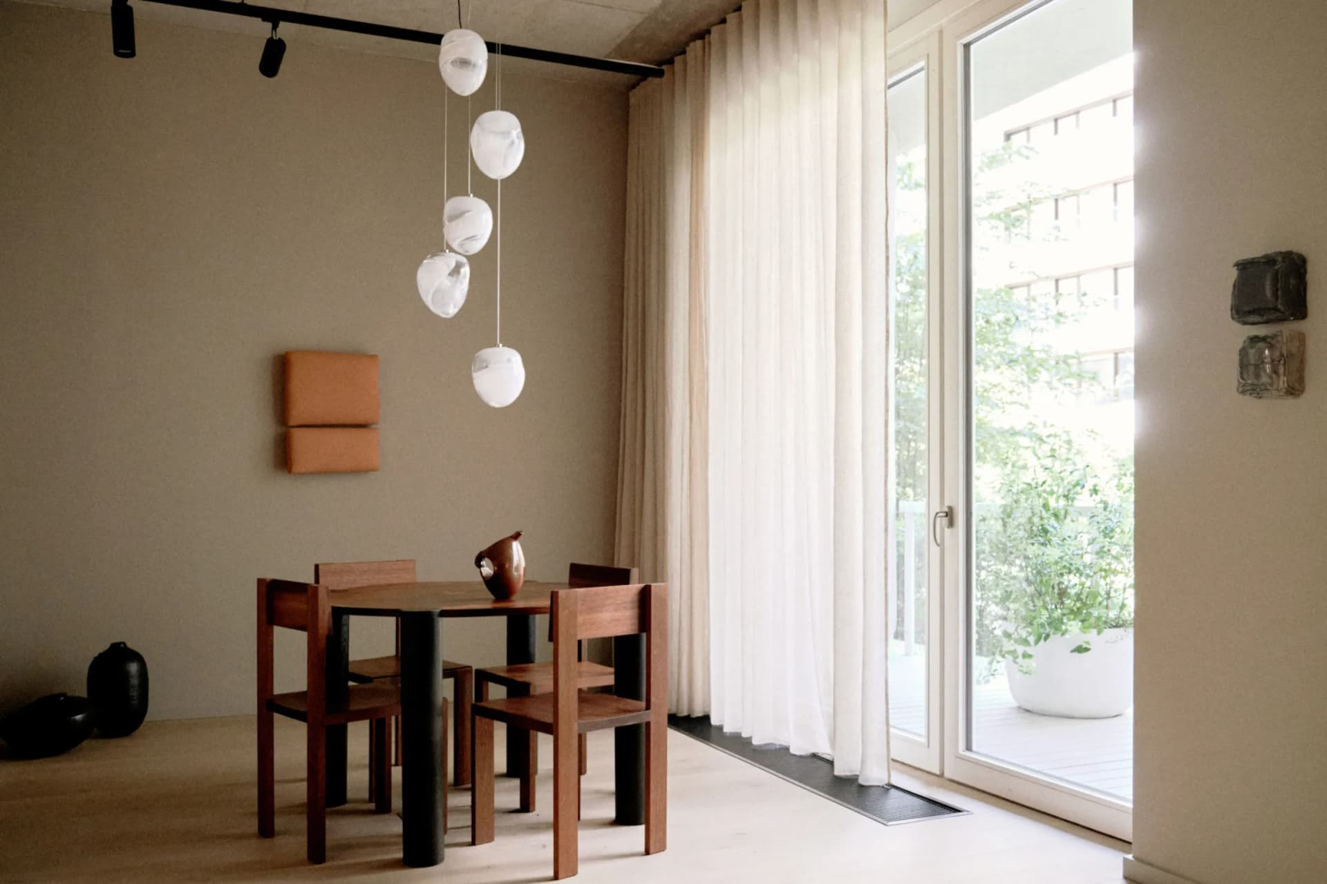 Salle à manger moderne avec une table et des chaises en bois, des suspensions blanches en forme de boules, des murs beiges agrémentés de coussins orange et de grandes portes vitrées donnant sur une terrasse ensoleillée.