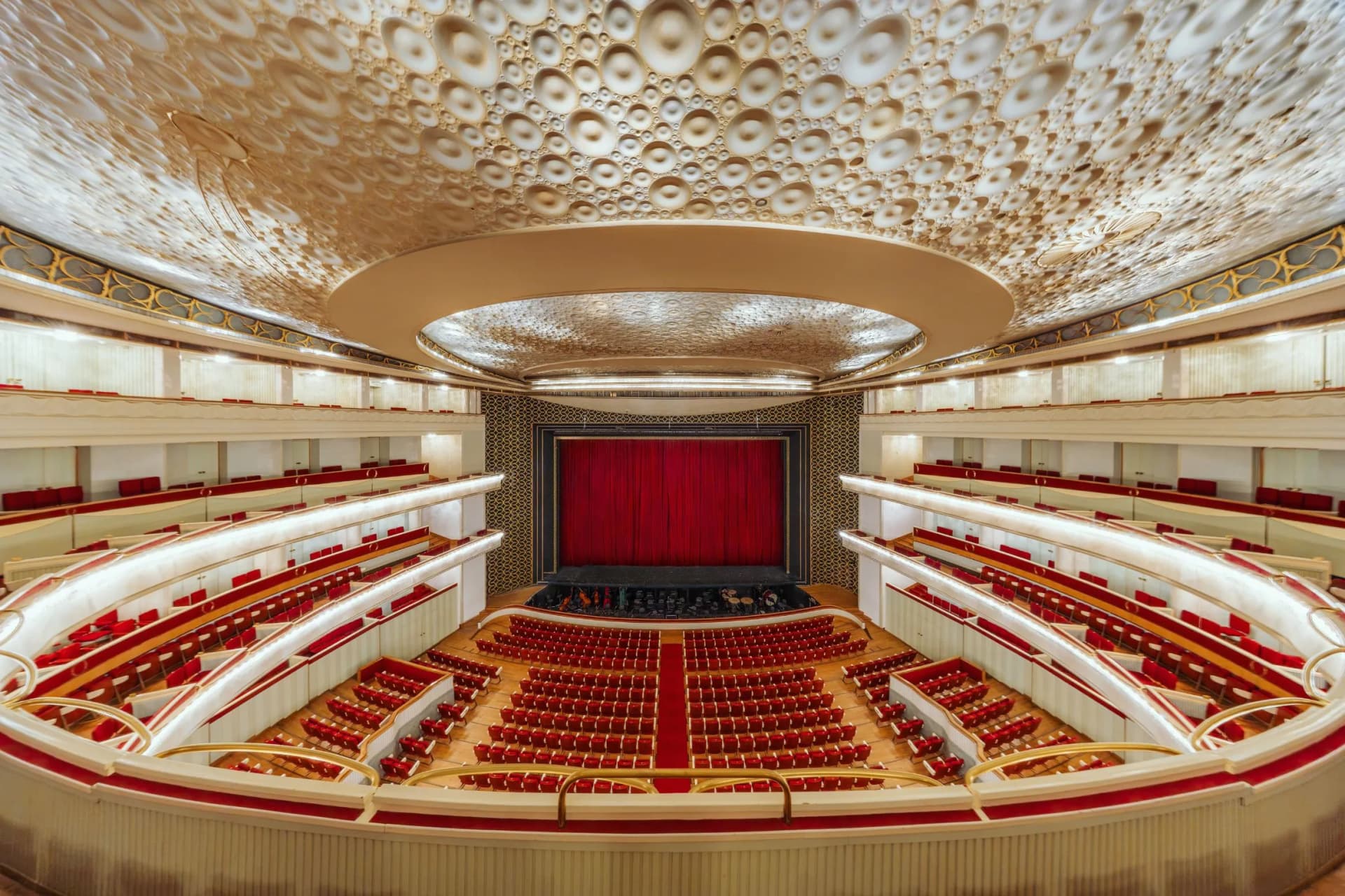 An opulent theatre interior featuring a ceiling with circular patterns, gilded balconies, red seats and a stage with red curtains.