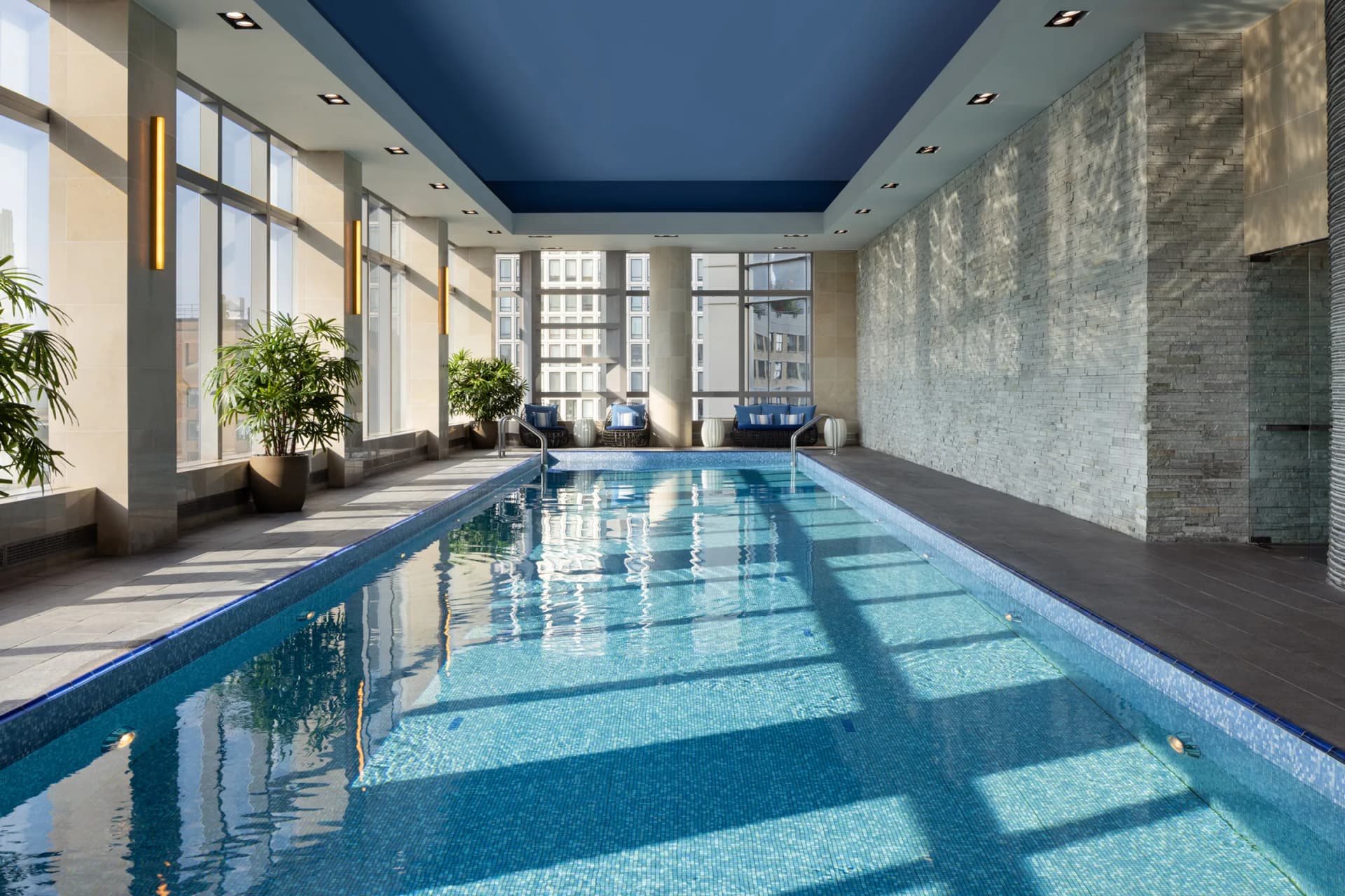 Indoor pool area in a modern space with a blue ceiling, stone walls, large windows, potted plants, and lounge seating along the pool.
