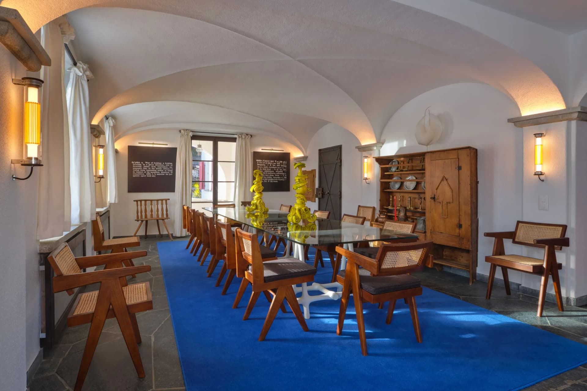 A room with a white vaulted ceiling, wooden chairs arranged around a glass table, a blue carpet, yellow sculptural table decorations and a wooden cupboard
